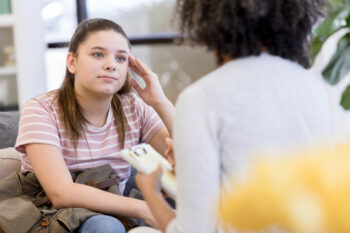 a teen girl sits on a couch facing a woman with a clipboard