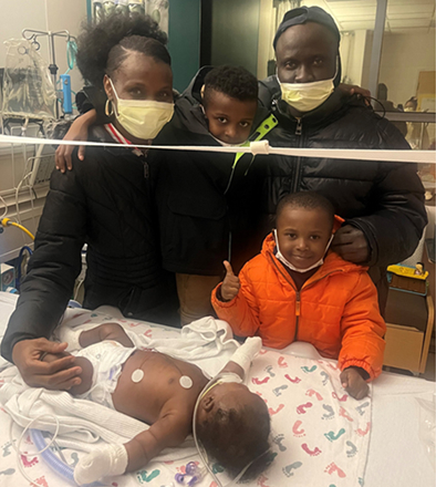 A family of four, wearing protective masks, stands together in a hospital room around a newborn baby lying in a medical crib.