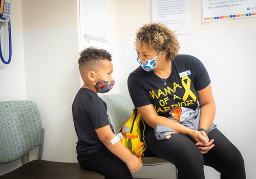 Kelli (right) and her son (left) sit in a medical office and are talking. They are both wearing masks, and Kelli is wearing a shirt that says &ldquo;Mama of a Warrior&rdquo; with the yellow cancer awareness ribbon.