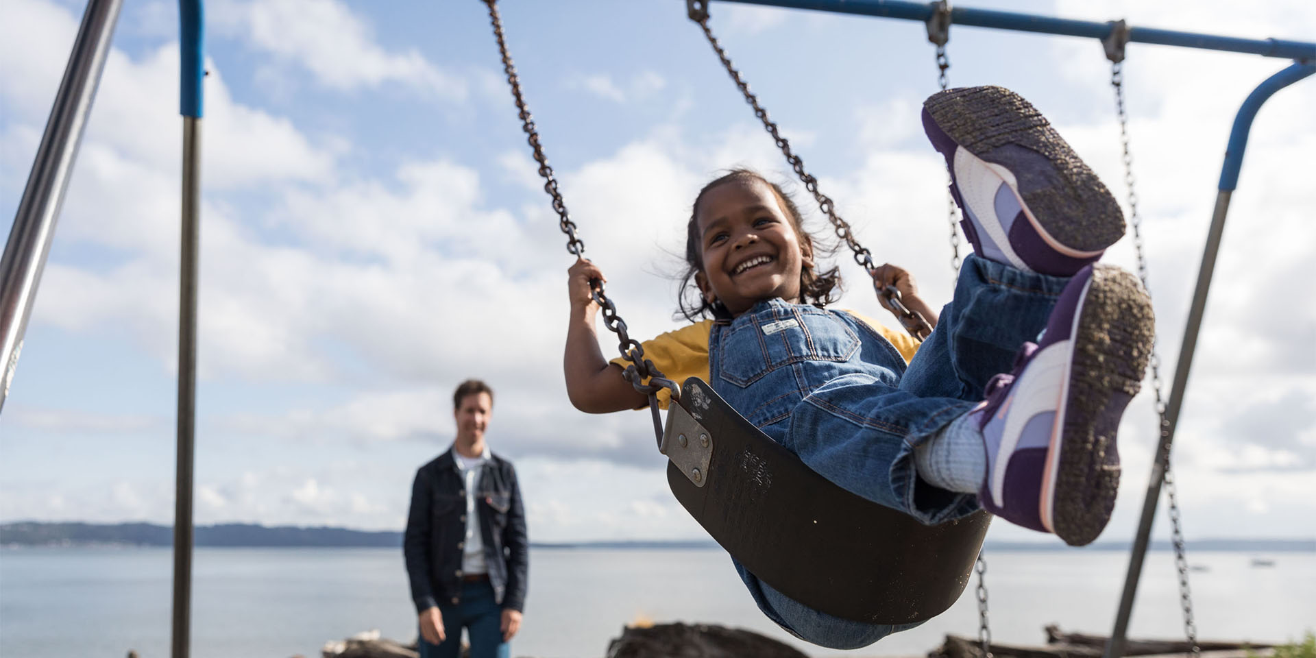 A school-aged girl with a giant smile on her face swinging on a swing as a man watches from the background