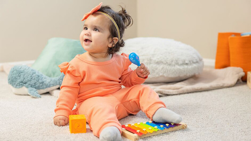 a smiling toddler girl with dark hair wearing coral colored sweatshirt holding a small blue maraca in her left hand 