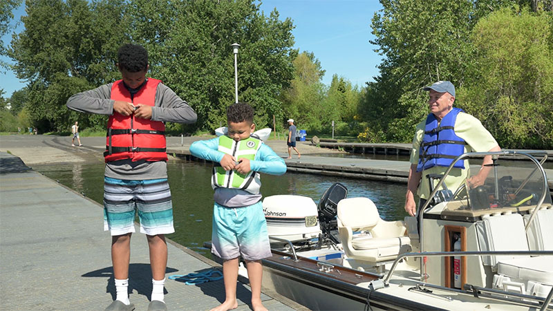 Two boys put on life jackets as a man in boat watches
