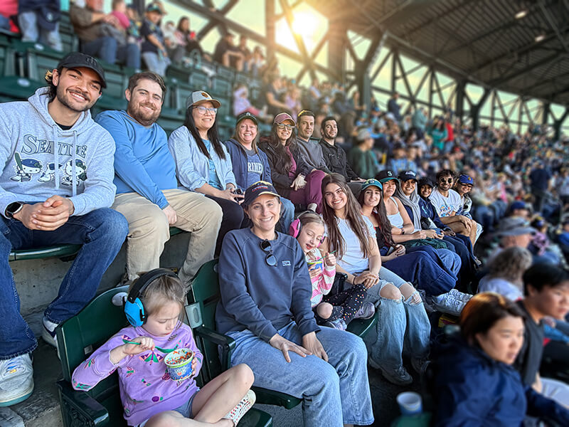 A large group of smiling people in the seats at T-Mobile Park during a Seattle Mariners baseball game.