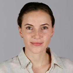a close up photo of a woman smiling at the camera. Her hair is dark brown and neatly tied up away from her face. She is wearing a tan, collared, button down shirt.