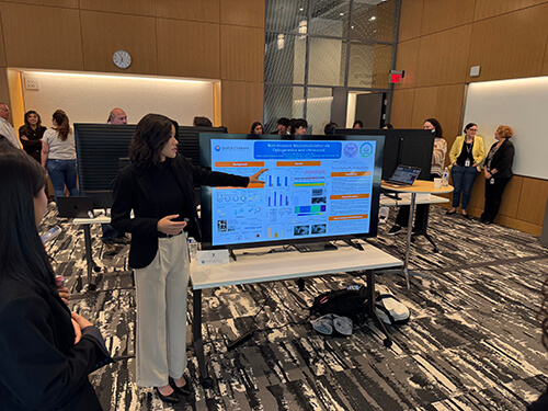 A student presenter stands beside a large display screen showing research charts and text, speaking to an audience in a modern indoor meeting space.
