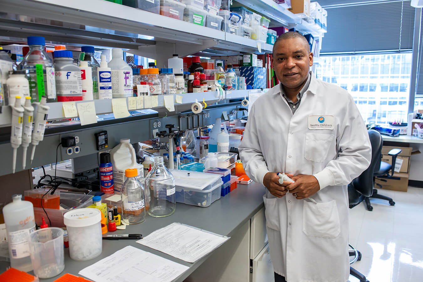 Researcher Franck Kalume, wearing a lab coat, stands in a busy laboratory setting, looking at the camera.