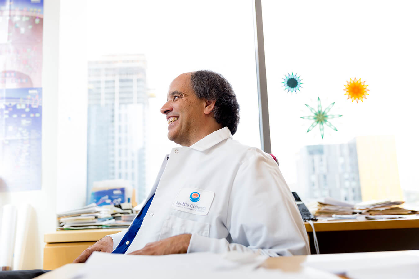 Dr. Nino Ramirez, wearing a lab coat, smiling in an office with city views and graphics of viruses.