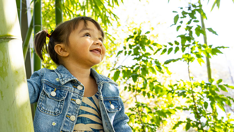 Pediatric patient in jean jacket stands among bamboo and smiles in the sunshine